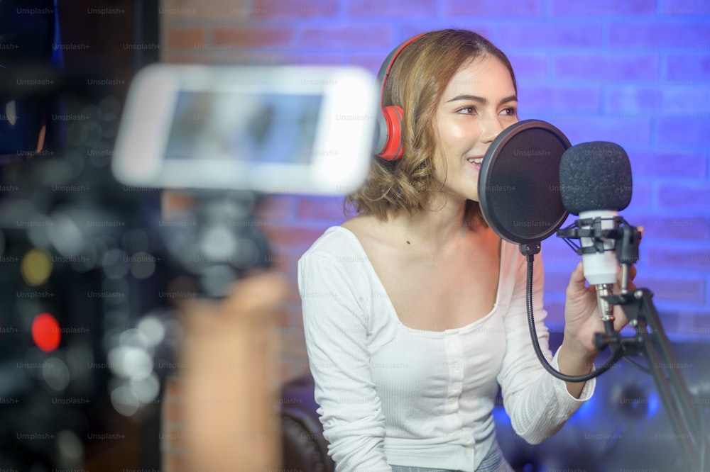 A young smiling female singer wearing headphones with a microphone while recording song in a music studio with colorful lights.