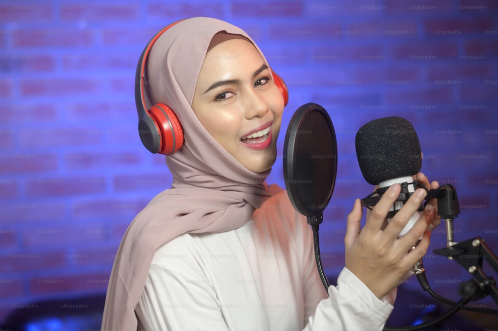 A young smiling muslim female singer wearing headphones with a microphone while recording song in a music studio with colorful lights.