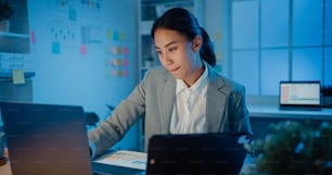 Young Asia cheerful professional woman sitting on desk focus concentrate working on laptop and digital tablet check detail final work in office at night. Lady business suit, Startup business concept.