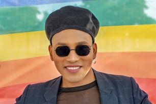 Young latin gay man with make-up on wearing a fashionable hat and sunglasses, isolated on a Rainbow flag background. LGBT.
