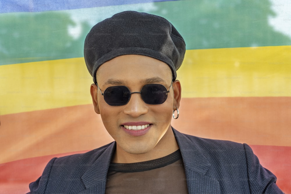 레인보우 깃발 배경에 고립된 세련된 모자와 선글라스를 쓰고 화장을 한 젊은 라틴 게이 남자. 성소수자.