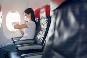 A traveling woman wearing protective mask onboard in the aircraft using smart watch, travel under Covid-19 pandemic, safety travels, social distancing protocol, New normal travel concept