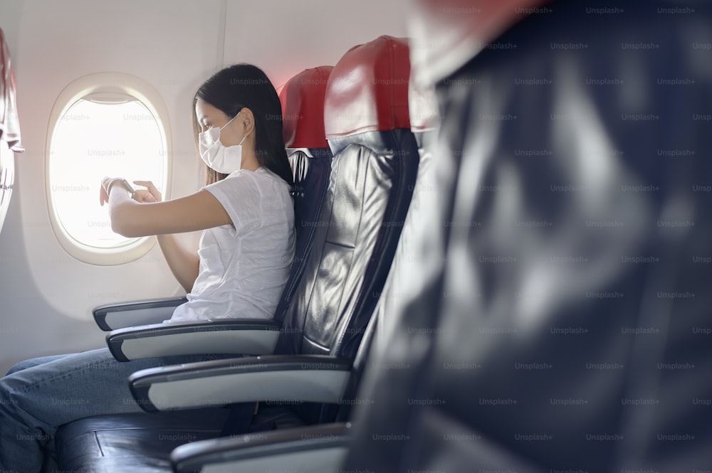 A traveling woman wearing protective mask onboard in the aircraft using smart watch, travel under Covid-19 pandemic, safety travels, social distancing protocol, New normal travel concept