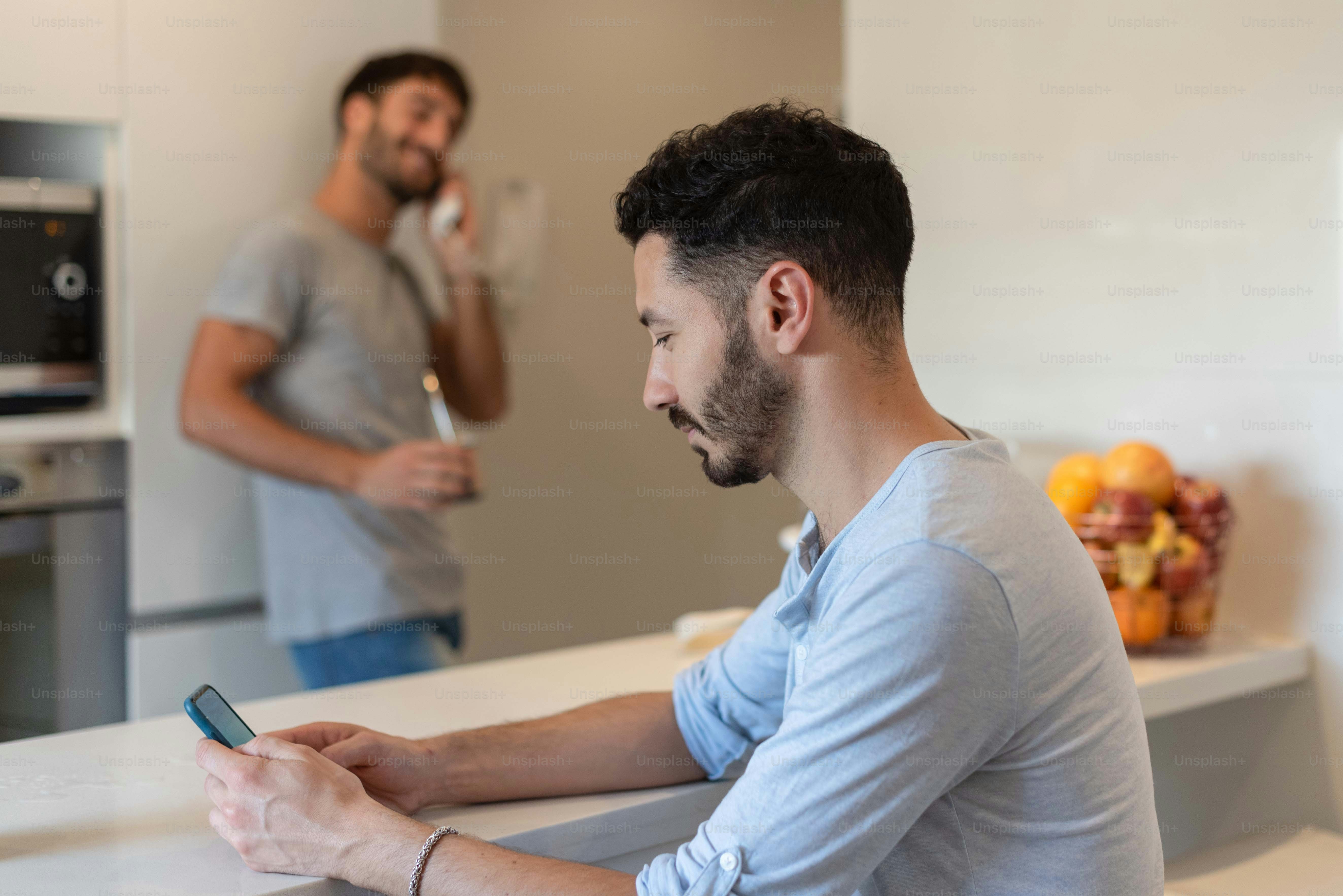 Young man using his smart phone while his boyfriend is answering a call at the door phone