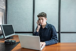 Joven trabajando en su computadora portátil mientras habla por teléfono en la oficina.