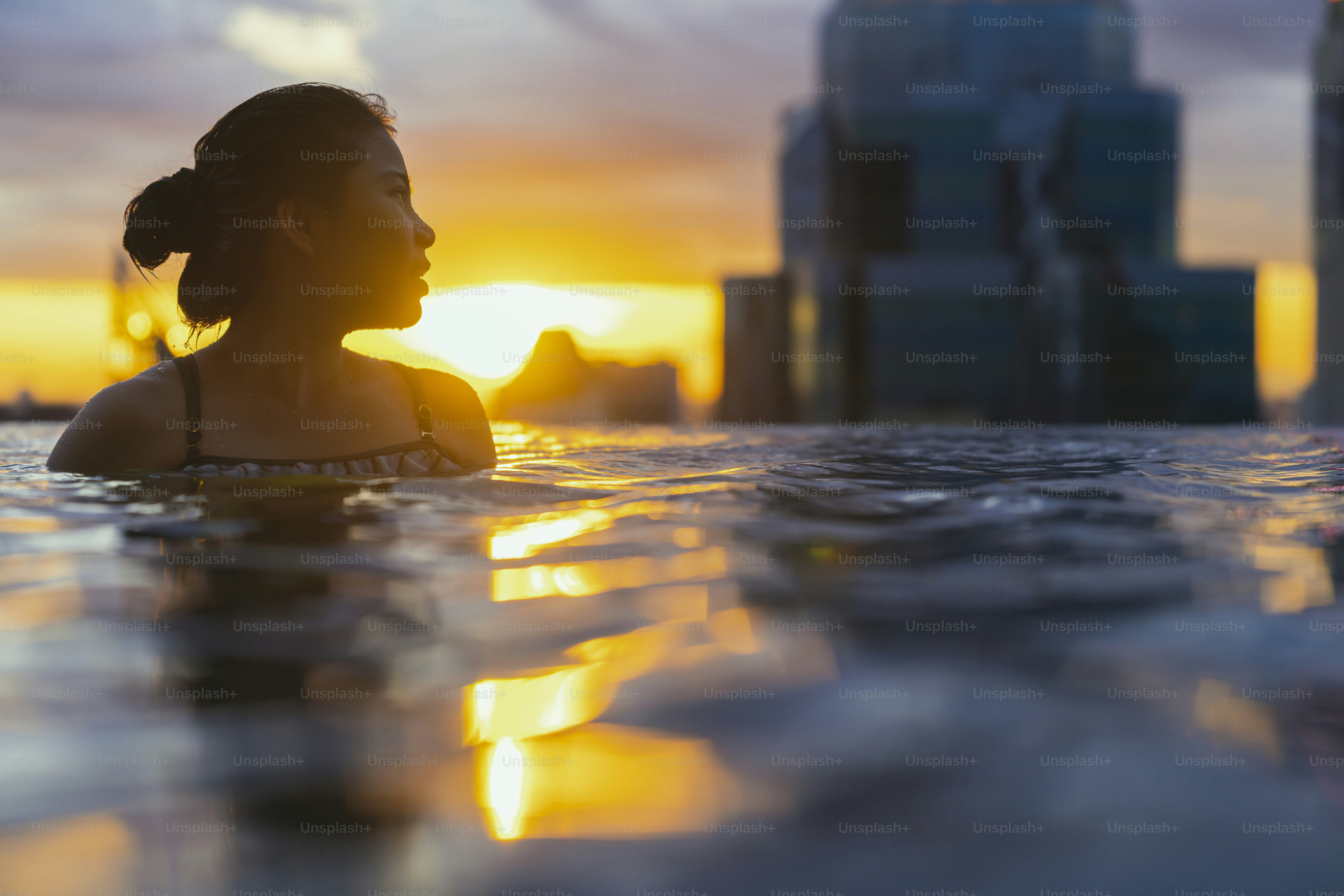 Siluet hitam wanita asia memercikkan air pada liburan musim panas bersantai di kolam renang tanpa batas dengan pemandangan matahari terbenam laut biru dengan skyscape bertingkat tinggi pusat kota Gaya hidup kebahagiaan sehat