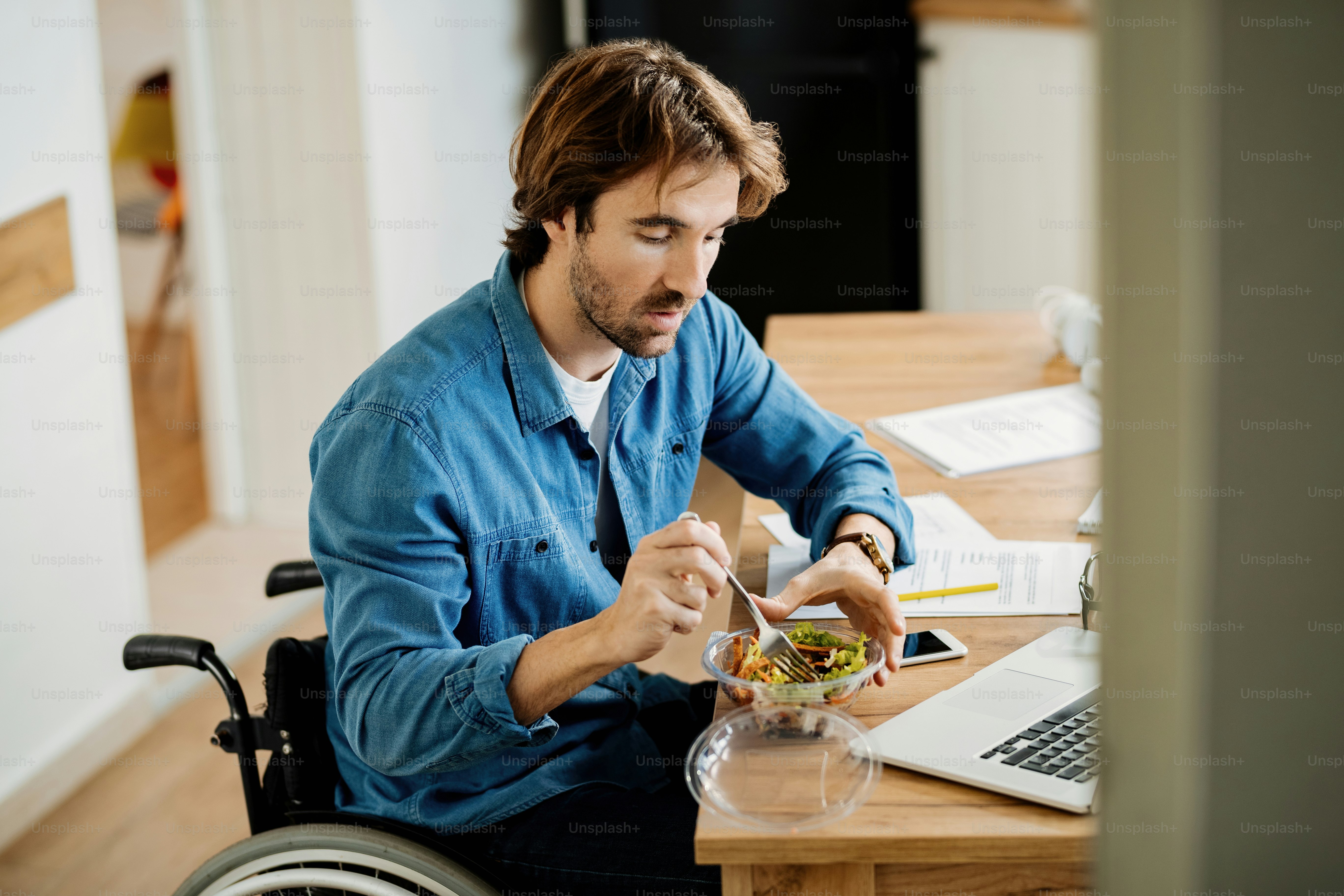 Young businessman in wheelchair working at home and eating salad on lunch break.