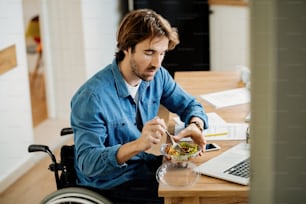 Young businessman in wheelchair working at home and eating salad on lunch break.