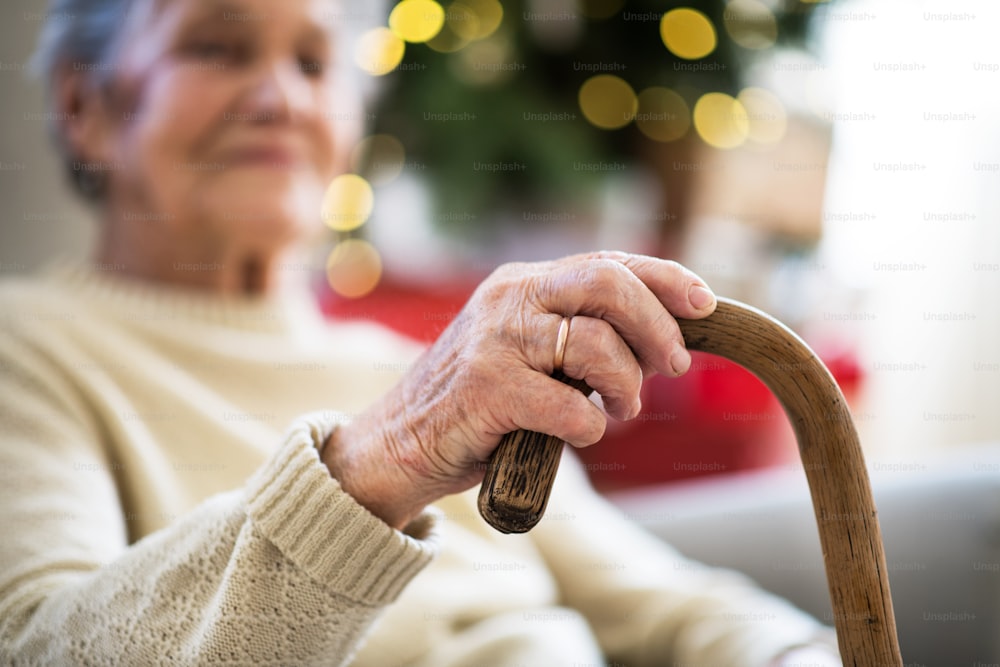 A close-up of a lonely senior woman sitting on an armchair at home at Christmas time, holding a walking stick.