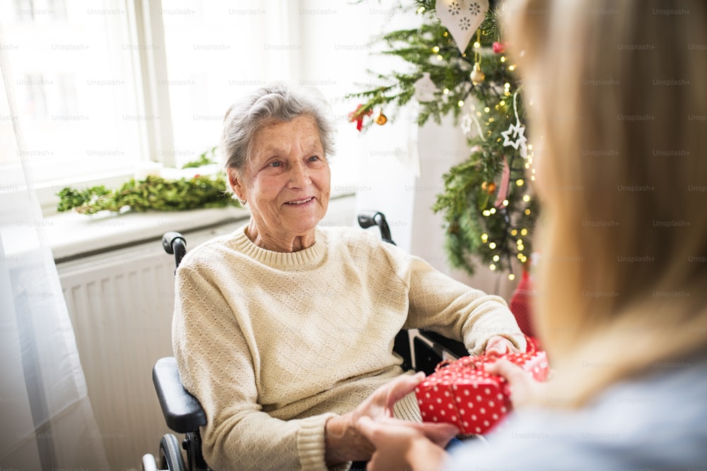 Un visitatore sanitario irriconoscibile e una donna anziana in sedia a rotelle con un regalo a casa nel periodo natalizio.