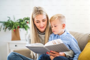 Una madre y un niño pequeño sentados en un sofá en casa en Navidad, leyendo un cuento de un libro.