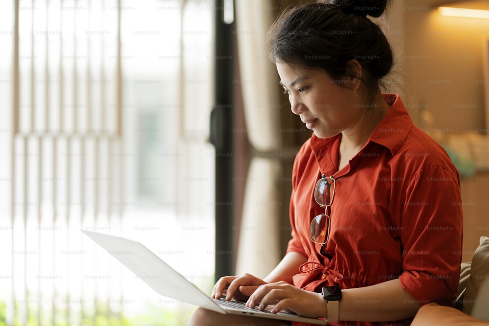 집에서 일하면서 거실에서 노트북을 사용하여 캐주얼하게 휴식을 취하고, 아시아 여성 성인 손 입력 검색 또는 노트북을 통한 문자 메시지 행복으로 집에서 일하는 평화로운 미소 아이디어 개념