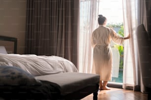 Junge asiatische Frau steht offen weiße Vorhänge am Fenster, am Morgen nach dem Aufwachen im Schlafzimmerhotel. Frau wachen mit einem frischen auf und öffnen die Vorhänge an den Fenstern.