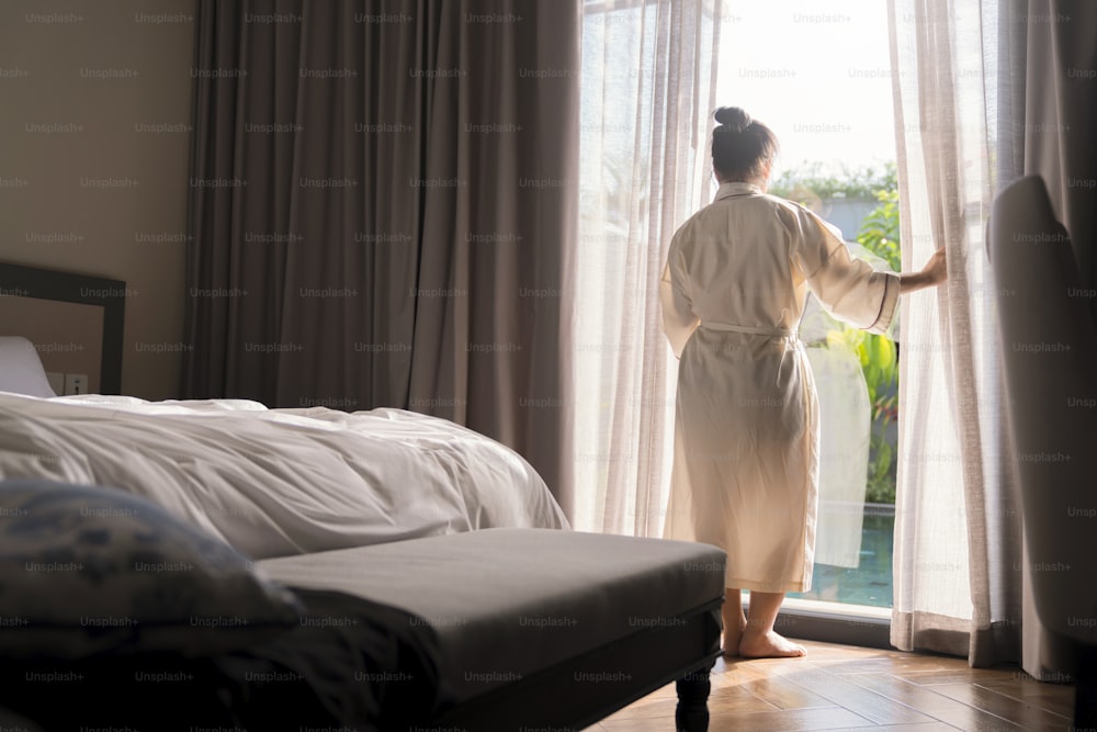 Giovane donna asiatica in piedi aperto tende bianche trasparenti alla finestra, la mattina dopo essersi svegliato nella camera da letto hotel. La donna si sveglia con un fresco e apre le tende alle finestre.