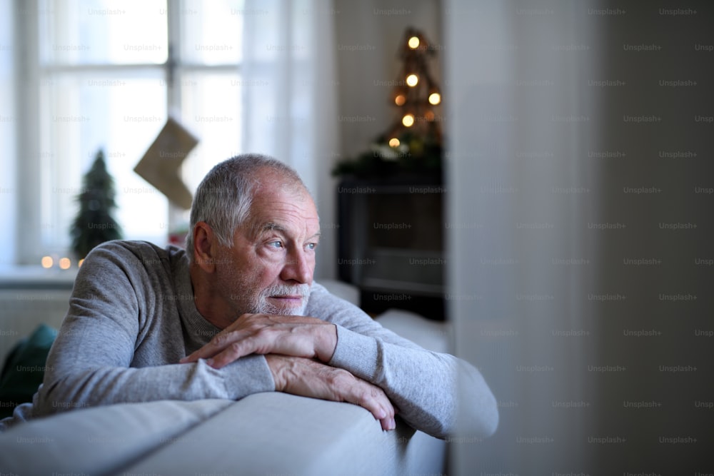 Portrait of lonely senior man sitting on sofa indoors at Christmas, solitude concept.