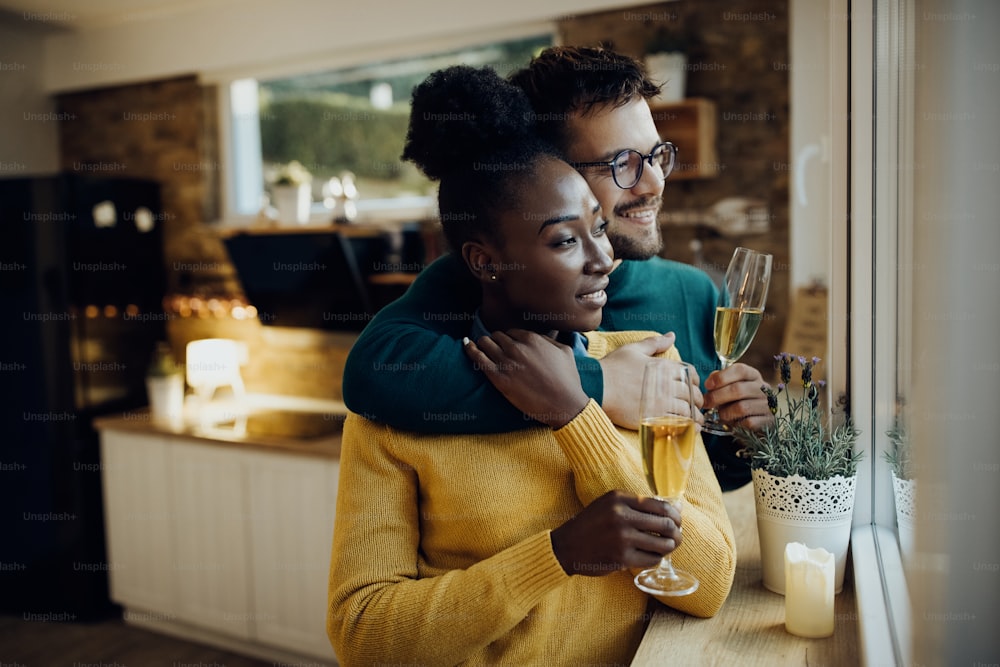 Glückliches gemischtrassiges Paar, das sich umarmt, während es zu Hause ein Glas Champagner am Fenster trinkt.