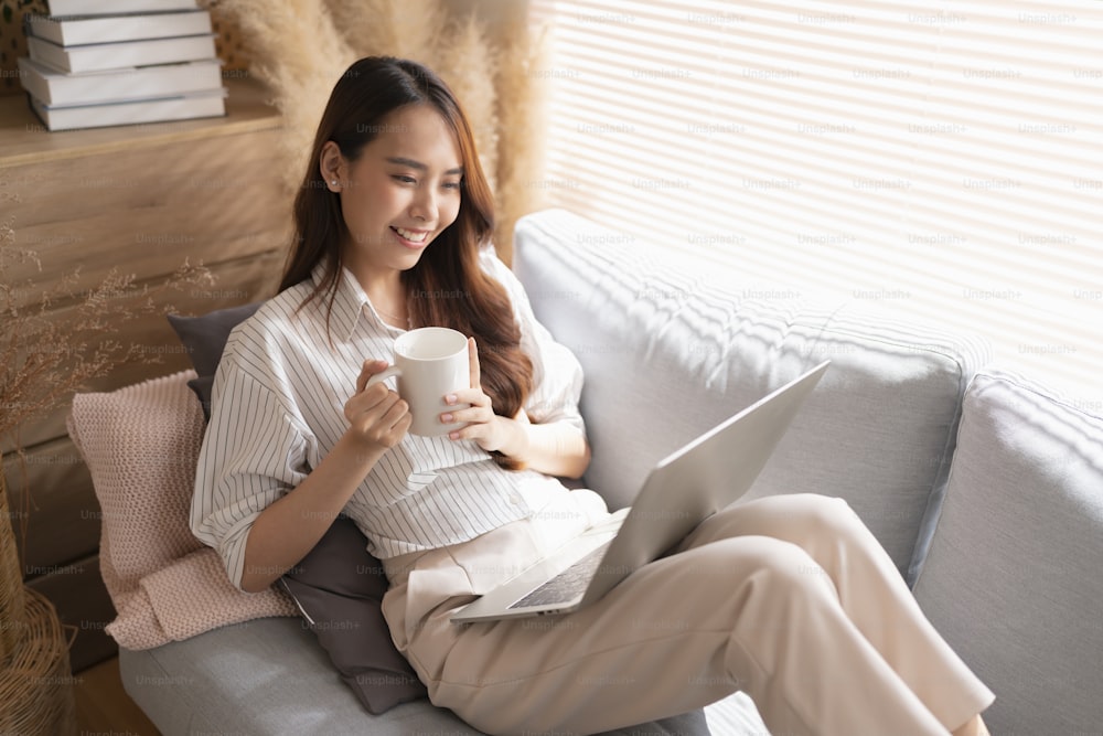 自宅で働いている若い魅力的なアジアの女性は、ラップトップチェック検索スケジュール作業プロセス計画レポートとレジャー朝の光でリビングルームのソファでリラックスし、平和な瞬間を過ごします
