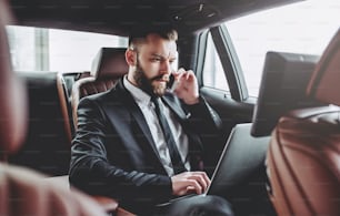 Young handsome businessman is sitting in luxury car. Serious bearded man in suit is working with laptop and talking on smart phone while being in trip.