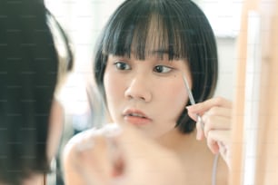 Quarantäne-Lifestyle-Konzept für den Aufenthalt zu Hause. Junge erwachsene asiatische Frau, die sich selbst schneidet, schlägt Haarschnitt mit einer Schere. Augen, die in einen Spiegel schauen. Hintergrund am Tag mit Naturlicht.