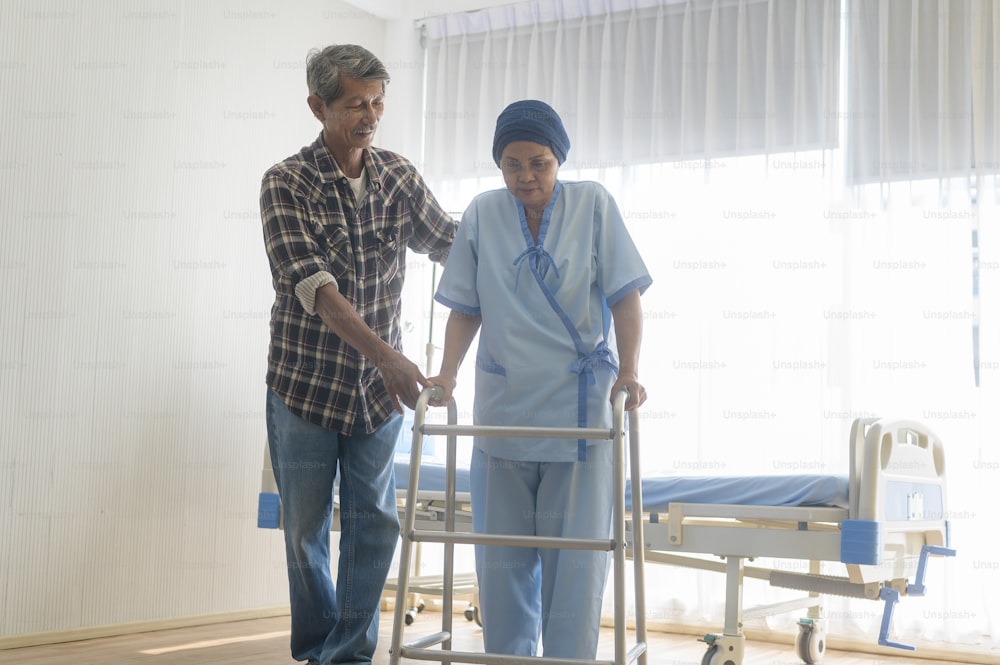 Un homme âgé aidant une patiente atteinte de cancer portant un foulard avec un déambulateur à l’hôpital, aux soins de santé et au concept médical