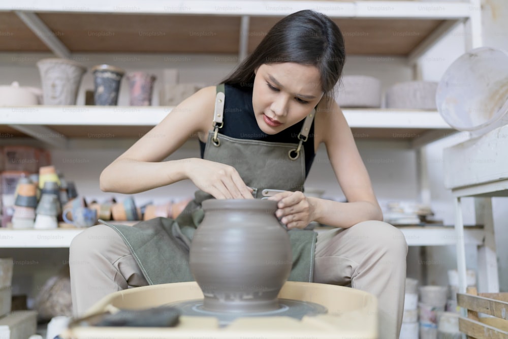 Close up de mãos femininas ceramist artista asiático que trabalham na roda dos oleiros, mulher da escultura feminina asiática com mãos sujas molhadas moldando vaso de argila na roda de cerâmica no estúdio da oficina