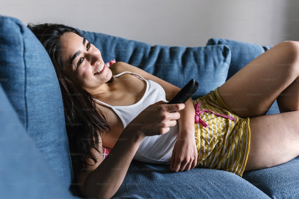 젊은 라틴 여자는 거실의 소파에 누워 있고 멕시코의 집에서 TV를 보고 있습니다.