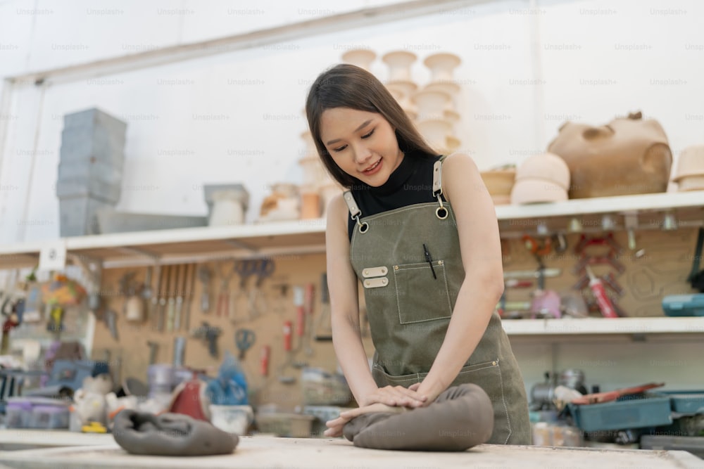 Giovane donna asiatica artista femminile indossare grembiule che fa vaso di argilla ciotola Bella giovane scultrice crea un vaso di arte di argilla nel magazzino dello studio di casa, ceramista artista mano femminile riscaldare massaggiare argilla sporco