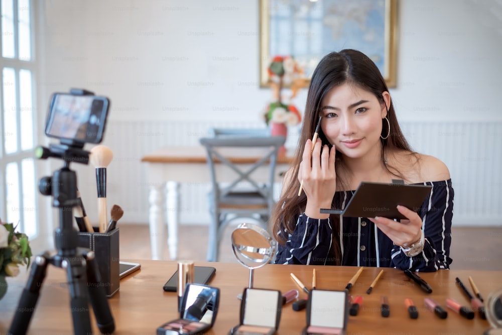 Joven bloguera asiática con maquillaje cosméticos grabando videoclip en línea por teléfono inteligente en casa. Enseñanza y venta de productos en línea.