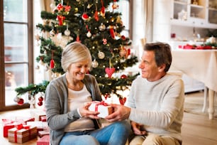 Seniorenpaar sitzt auf dem Boden vor dem beleuchteten Weihnachtsbaum in ihrem Haus und beschenkt sich gegenseitig.