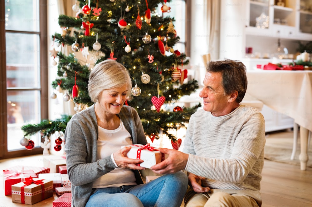 Senior couple sitting on the floor in front of illuminated Christmas tree inside their house giving presents to each other.
