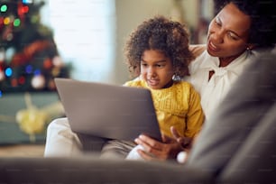 Happy African American little girl and her mother surfing the net on laptop while relaxing at home on Christmas.