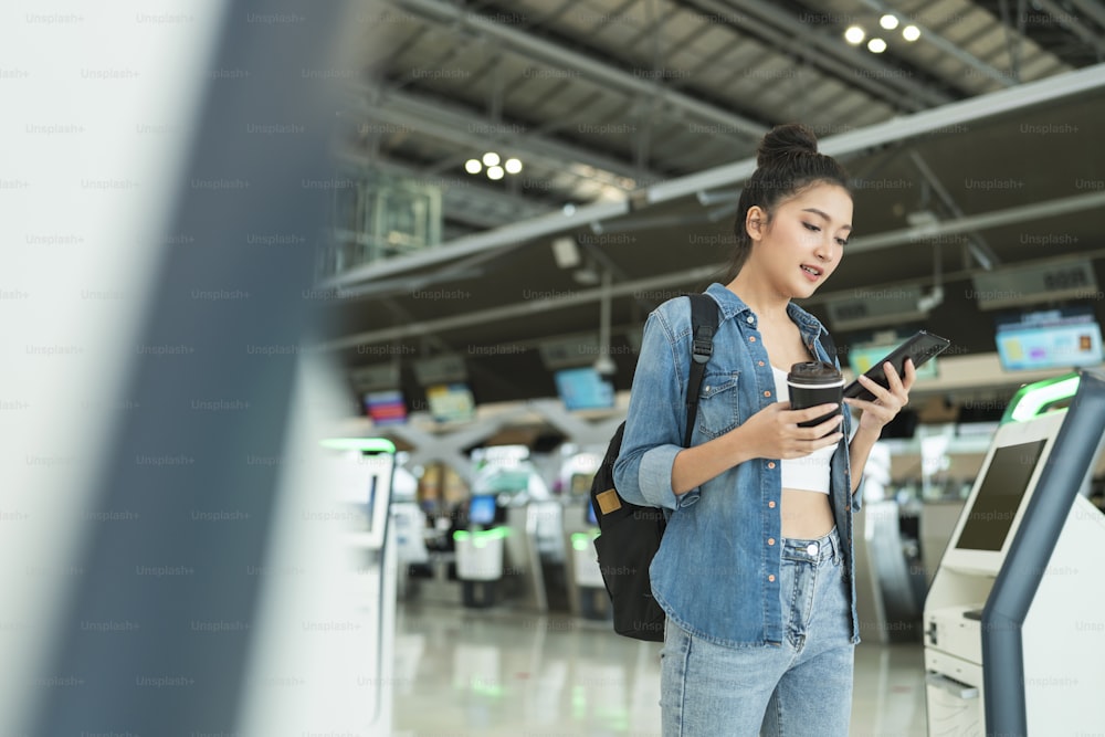 expatのデジタル遊牧民の旅行と仕事、アジアの陽気な女性のカジュアルな布は、空港ターミナル旅行のコンセプトでスマートフォン予約チェックインフライトオンラインチケット情報アプリケーションを使用しながら歩いています