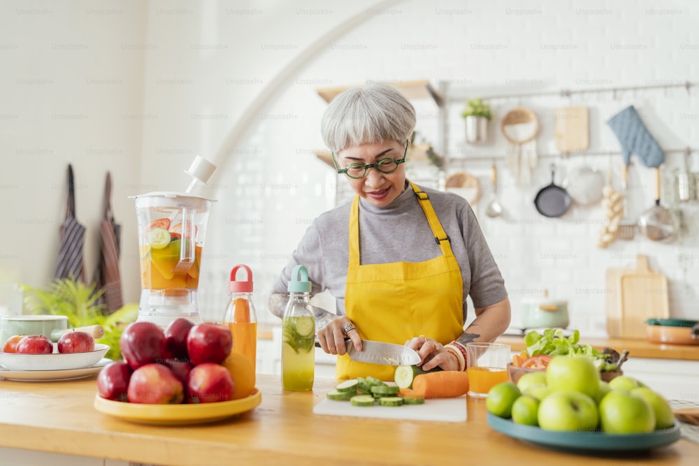 Reife lächelnde Tattoo-Frau, die Salat, Obst und Gemüse isst. Attraktive reife Frau mit frischem grünem Obstsalat zu Hause. Seniorinnenschürze steht in der Küchentheke und entspannt sich im Haus