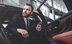 Young handsome businessman is sitting in luxury car. Serious bearded man in suit with smart phone in hands.
