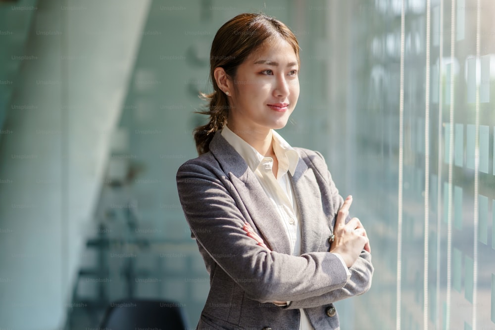 Hermosa mujer de negocios asiática sonriente de pie con los brazos cruzados en la oficina mirando por la ventana.