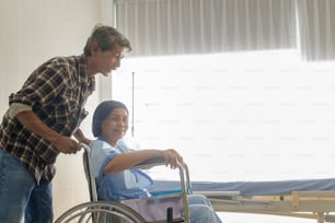 Un homme âgé aidant une patiente atteinte d’un cancer portant un foulard à se déplacer vers des fauteuils roulants à l’hôpital, aux soins de santé et au concept médical