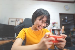 オタクスタイルのヤングアダルトアジアンゲーマーの女性は眼鏡をかけ、ハンドヘルドオンラインゲームをプレイします。勝利ムードを競う。人々は自宅で余暇のライフスタイルをします。