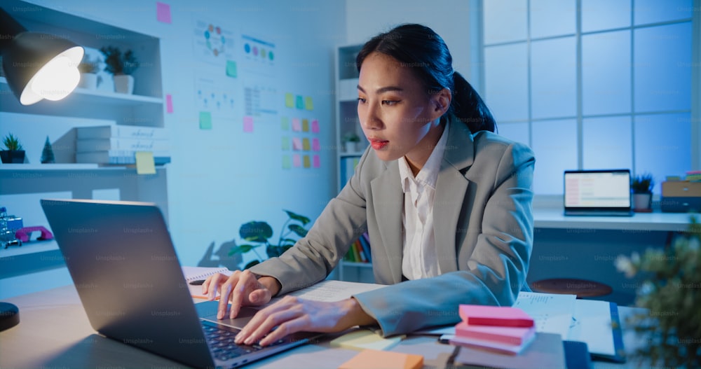 Young Asia cheerful professional businesswoman sitting on desk with laptop computer work online marketing in modern office at night. A woman in a business suit, Work and technology start up concept.