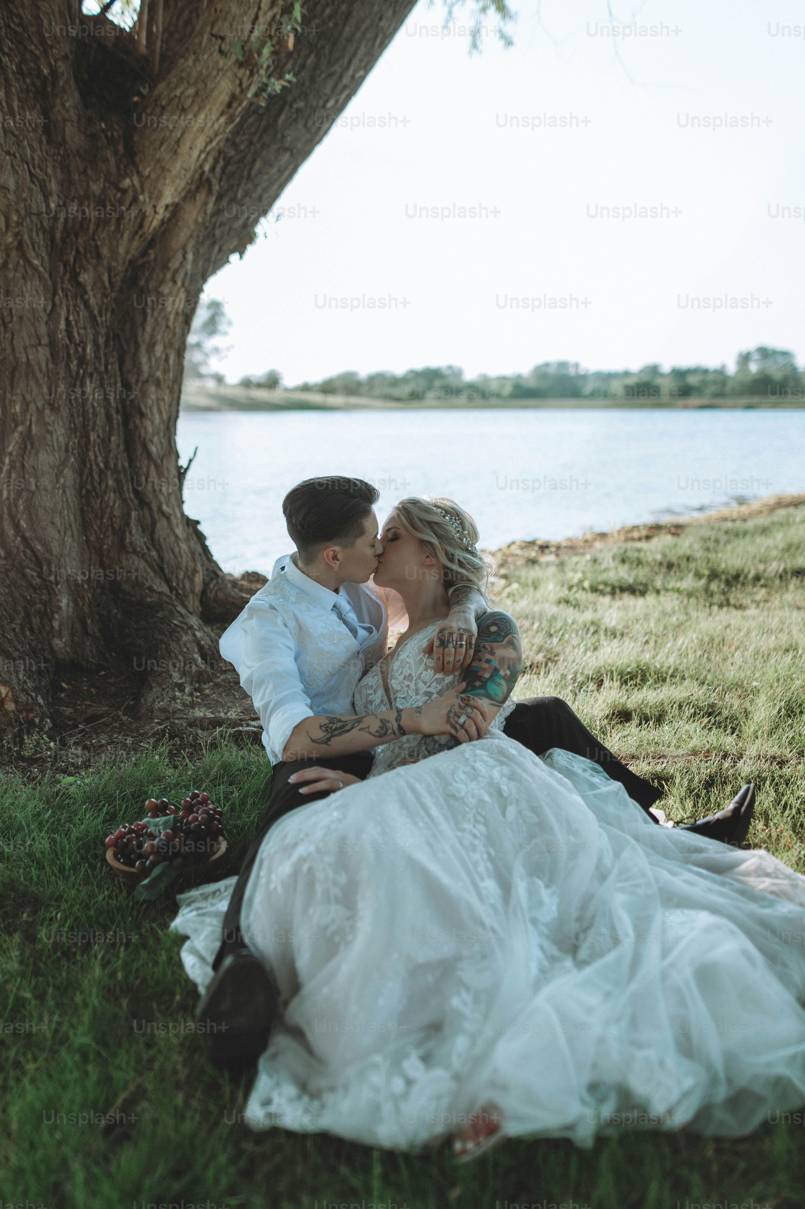 a bride and groom sitting under a tree by the water