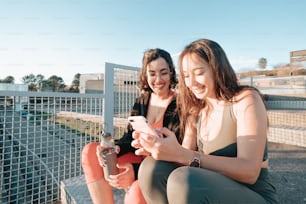 two women sitting on a fence looking at a cell phone