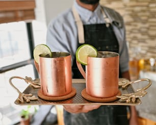 a man holding a tray with two copper mugs on it