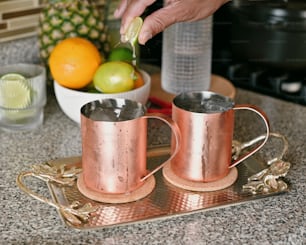 a pair of copper mugs on a tray with fruit in the background