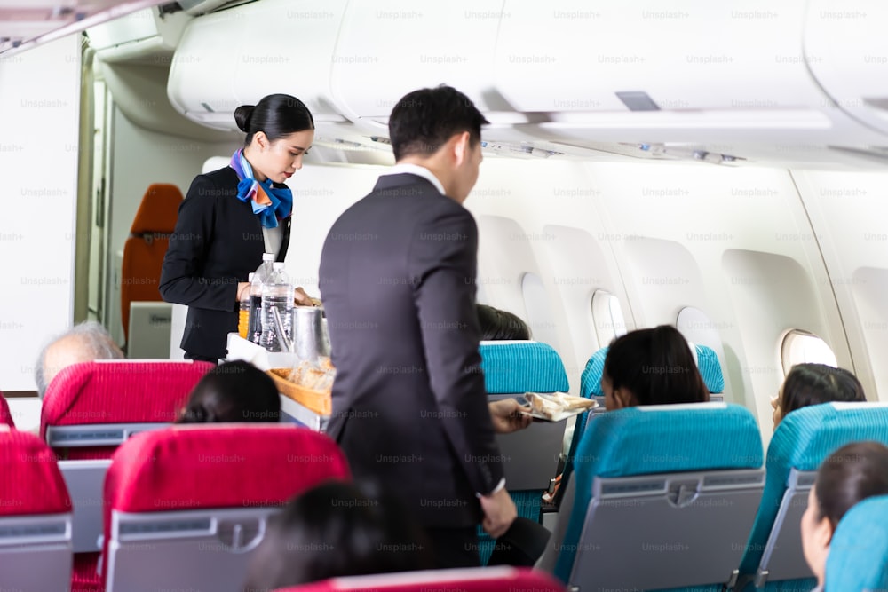 Assistente di volo asiatico femminile e maschile che serve cibo e bevande ai passeggeri sull'aereo. L'equipaggio di cabina spinge il carrello sul corridoio per servire il cliente. Concetto di lavoro e occupazione del servizio di compagnia aerea.