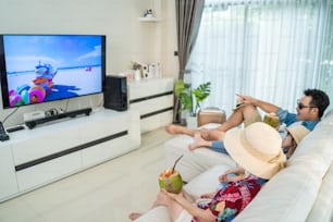 Asian Family watch TV screen, pretend on beach during summer in house. Happy Traveller people having fun stay at home, Parent spend time with young children girl in living room due to Covid19 lockdown