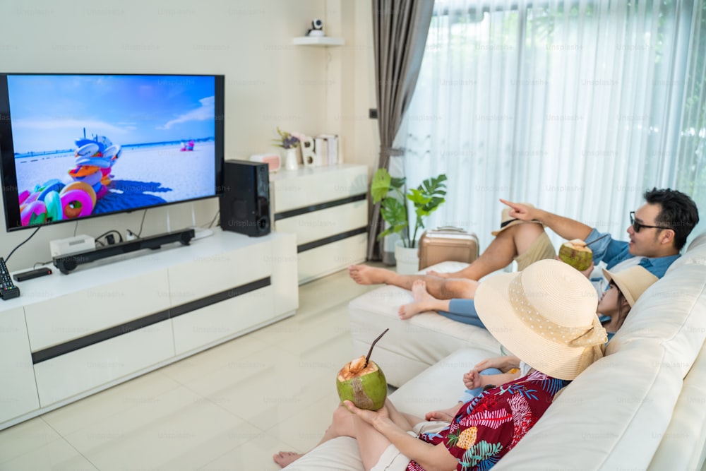 アジアの家族はテレビ画面を見て、夏の間は家でビーチでふりをします。楽しい時間を過ごしているハッピートラベラーの人々は家にいて、親はCovid19のロックダウンのためにリビングルームで幼い子供の女の子と時間を過ごします