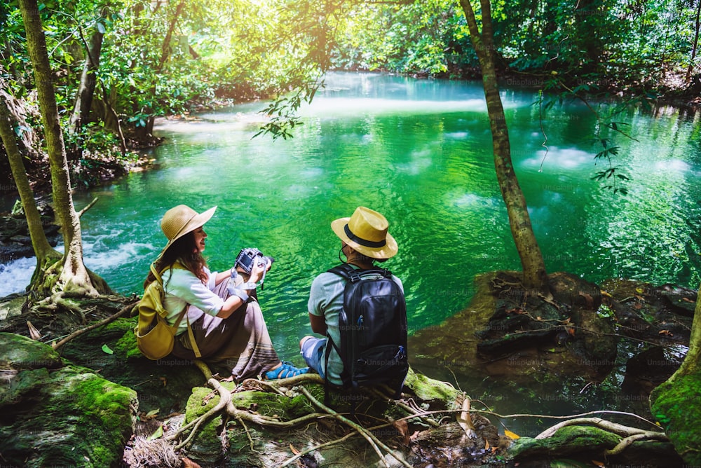旅行者、バックパックを背負ったカップル、岩に座ってリラックスしてください。緑のジャングルで自然を旅し、滝の景色を楽しんでいます。観光、ハイキング、自然研究。旅行中のカップル、休日に自然の写真を撮る。エメラルドストリーム。