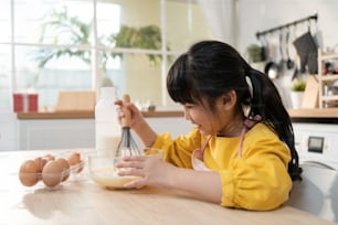 Retrato de una niña linda joven asiática haciendo panadería casera en la cocina. Adorable niño pequeño se sienta en la mesa sintiéndose feliz y disfruta aprendiendo a cocinar alimentos u horneando amasa masa de levadura con las manos en el hogar