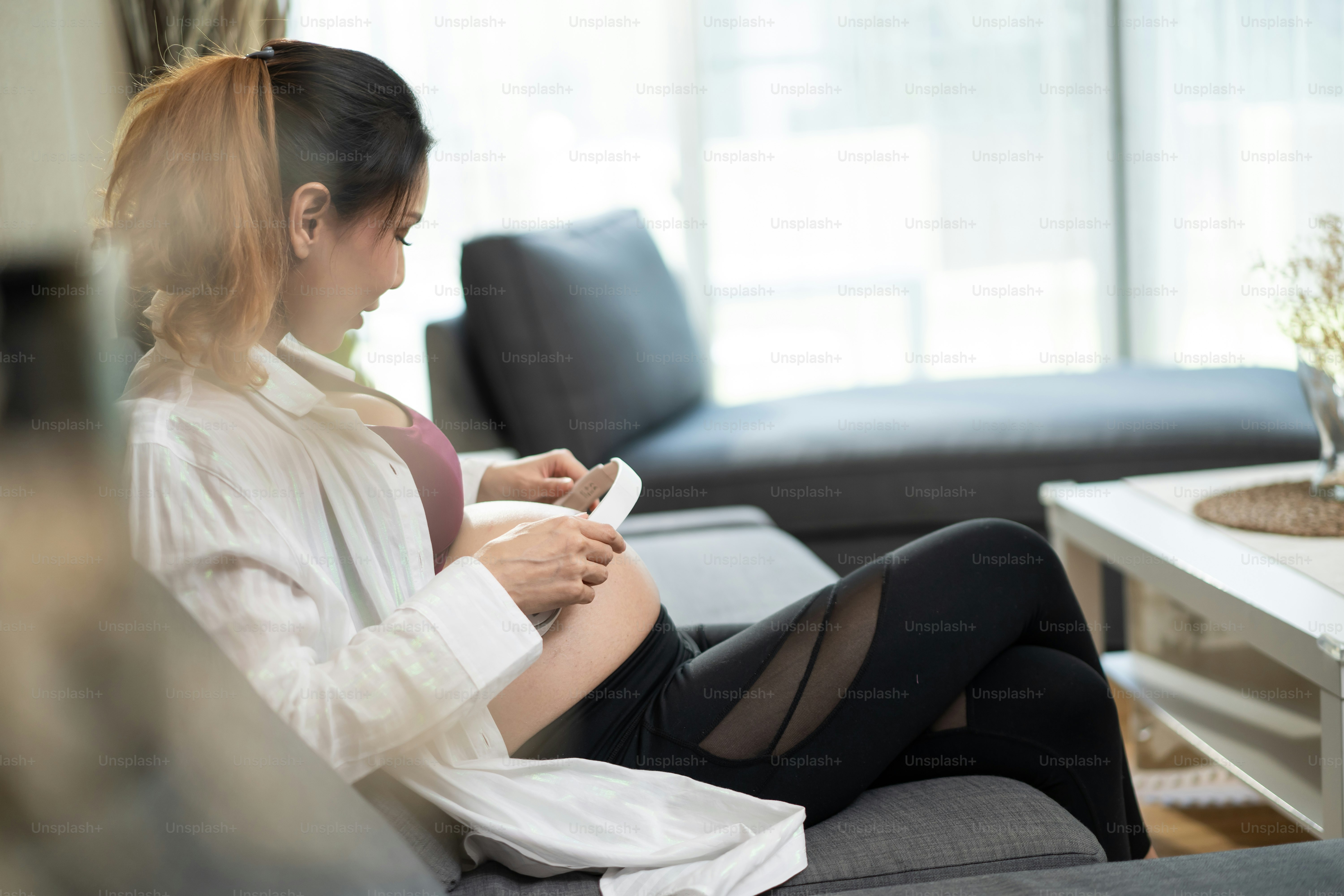 Asian young beautiful pregnant woman applying headphones on her belly. Attractive expectant pregnancy girl mother sit on sofa indoors in living room and play music for newborn baby in belly to listen.