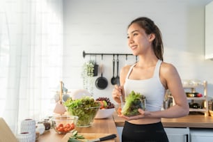 Asian attractive sport woman holding salad bowl and eat vegetables. Beautiful sport girl in sportswear enjoy eat clean vegetables after exercise for health in house. Diet and Healthy food concept.
