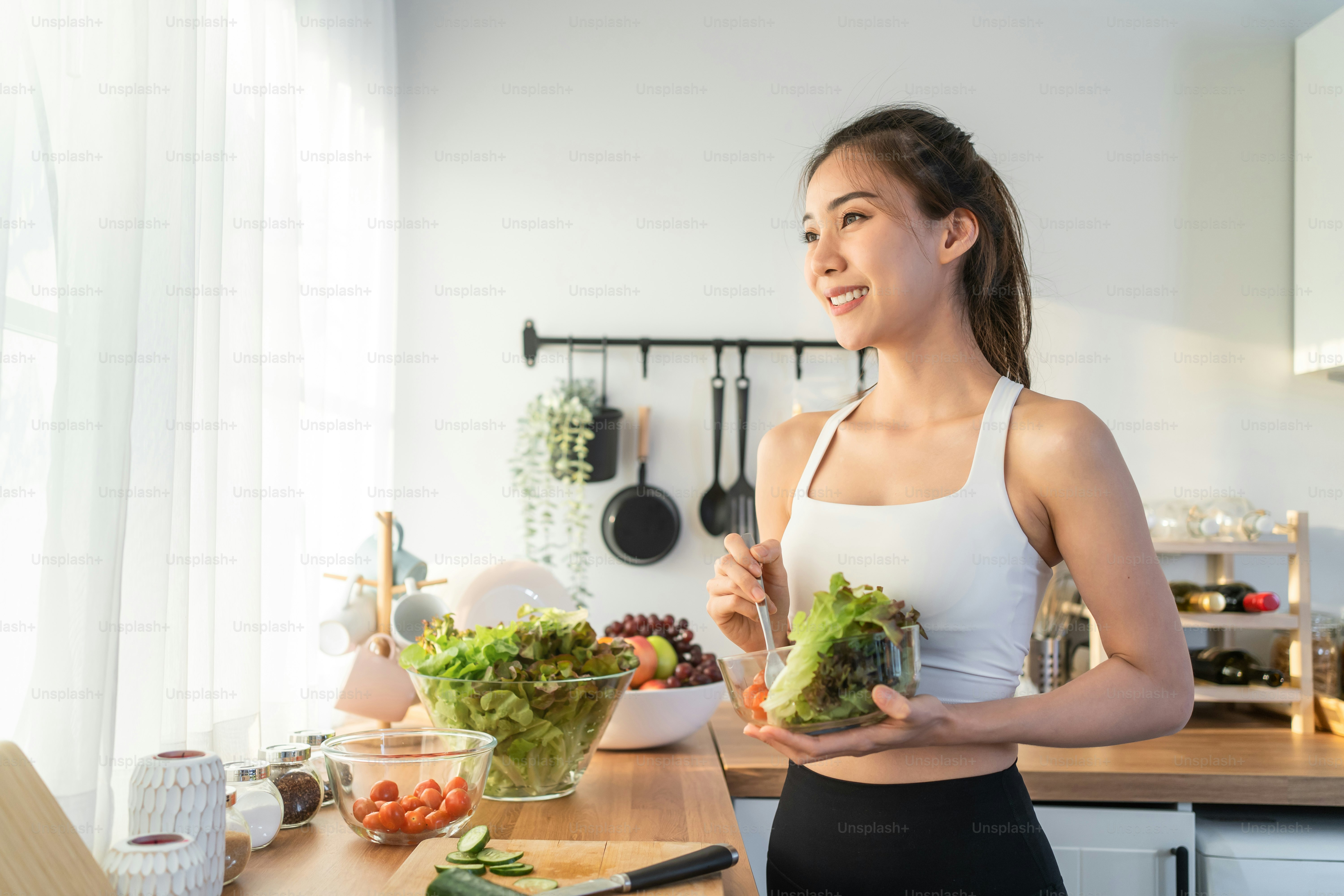 Asian attractive sport woman holding salad bowl and eat vegetables. Beautiful sport girl in sportswear enjoy eat clean vegetables after exercise for health in house. Diet and Healthy food concept.